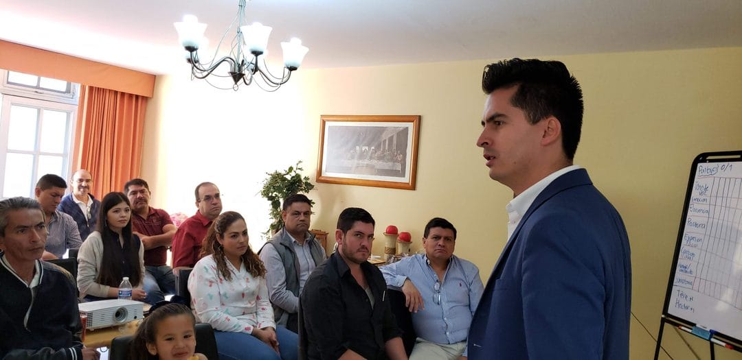 Poncho, de traje azul y camisa blanca, habla de pie frente a un grupo de unas 13 personas sentadas en una sala con candil, un cuadro…